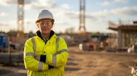 Confident female construction worker in safety gear standing at building site with cranes and equipment in background during golden hour sunset.