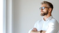 Confident Caucasian male professional with beard and glasses in white shirt standing by window with arms crossed, looking up thoughtfully in modern office space.
