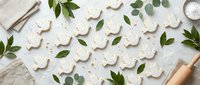 White gingerbread cookies shaped like reindeer arranged with fresh green leaves and baking tools on textured surface for holiday baking and festive cooking.
