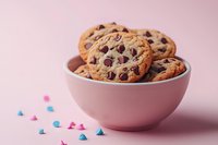 Fresh chocolate chip cookies in pink ceramic bowl against pastel background with scattered colorful sugar decorations, food photography for bakery marketing.