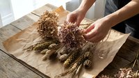 Hands arranging dried wheat, grains and natural plant stems on kraft paper for craft, decoration or agricultural design projects.