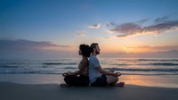 Two people practicing meditation together on beach at sunset with golden sky and ocean waves creating peaceful wellness moment for mindfulness and relaxation.