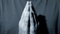 Elderly hands pressed together in prayer position showing wrinkled skin and age spots against dark fabric background conveying spirituality and devotion.