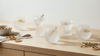 Modern glass tea set with transparent teapot and cups arranged on wooden table with dried herbs and spoons for brewing ceremony and relaxation.