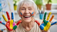 Joyful senior Caucasian woman with colorful paint on hands and face smiling in art therapy session, expressing creativity and happiness in retirement.