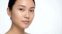 Young Asian woman with clear, glowing skin and natural makeup looking at camera against white background for skincare and beauty advertising.