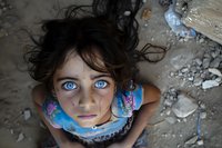 Young girl with striking blue eyes gazes upward intensely, wearing floral dress against rough concrete background, dramatic portrait emphasizing innocence and emotion.