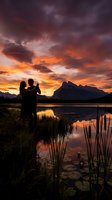 Romantic couple silhouette embracing at mountain lake during dramatic sunset with vibrant orange and purple sky reflection in calm water.
