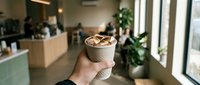 Hand holding takeaway coffee cup with latte art in modern cafe interior with natural lighting and plants creating cozy atmosphere for coffee lovers.