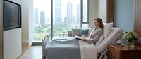 Professional businesswoman relaxing in modern hospital bed with city skyline view through large windows, representing healthcare comfort and recovery.