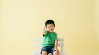 Young Asian boy, approximately 2-3 years old, wearing green t-shirt and denim shorts, sitting on white chair against yellow background, smiling and giving thumbs up gesture for childcare, parenting, and happiness concepts.