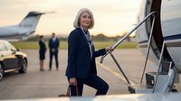 Mature woman with blonde hair, professional appearance, wearing dark business suit and holding briefcase, ascending airplane stairs at private airfield during golden hour with luxury vehicles and aircraft in background.