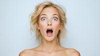 Surprised young Caucasian woman with blonde messy hair and natural makeup expressing shock with wide open mouth and eyes against light background.