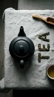 Black ceramic teapot on linen cloth with loose green tea leaves arranged as letters, wooden scoop, and tea cup in moody flat lay.