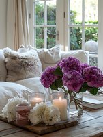 Cozy home interior with purple hydrangeas in vase, lit candles on wooden tray, and white cushions by bright window. Soft natural lighting creates peaceful atmosphere.
