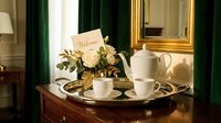 Elegant hotel room welcome tray with white tea set, fresh roses, and a greeting card on a wooden dresser with gold mirror and green drapes.