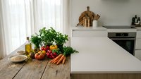Modern kitchen interior with fresh vegetables and herbs on wooden table, white countertops and built-in appliances creating clean minimalist cooking space.