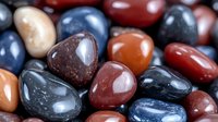 Polished gemstones in various colors including red jasper, blue sodalite, and orange carnelian create natural pattern of smooth tumbled stones with glossy surface.