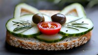Fresh cucumber cat face sandwich on seeded bread with cherry tomato nose, olive eyes and rosemary whiskers, creative food art for children's lunch or party snacks.
