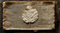 Single dried leaf on natural burlap textile with frayed edges against dark wooden background, creating rustic minimalist composition for nature-inspired decor.