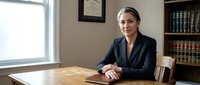 Female attorney with gray hair in professional dark blazer seated at wooden desk in law office with bookshelf background, suitable for legal services marketing.