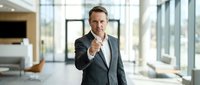 Confident middle-aged Caucasian businessman in dark suit pointing finger at camera in modern office lobby with large windows and contemporary furniture.