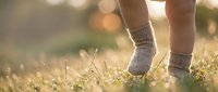 Child feet in cozy knit socks walking on grass during golden hour sunset with warm bokeh lights creating peaceful outdoor moment.