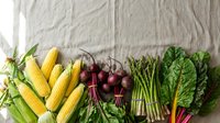 Fresh organic vegetables including corn, radishes, asparagus and leafy greens arranged on linen fabric for healthy cooking and farm-to-table cuisine concepts.