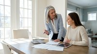 Professional mature blonde businesswoman mentoring young brunette colleague at modern office desk with laptop and documents in bright workspace.