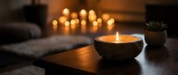 Cozy living room interior with burning candle in wooden bowl on coffee table, warm ambient lighting from blurred candles in background creating relaxing atmosphere.