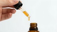 Hand holding dropper dispensing golden oil into amber glass bottle against white background for skincare and wellness applications.