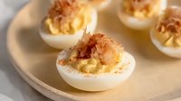 Creamy deviled eggs topped with crispy bacon bits and paprika spice on white ceramic plate, appetizing party snack photographed in soft natural light.