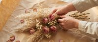 Hands arranging dried flowers and wheat stems on kraft paper, creating a rustic floral arrangement for wedding decoration or craft projects.