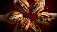 Hands holding variety of gourmet sandwiches in circle formation on dark background. Fresh deli meats, vegetables and bread for food photography and menu design.