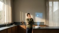 Young professional man in green sweater talking on phone in modern kitchen with natural sunlight streaming through windows and warm interior design.