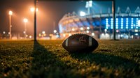 Football on grass field at night with stadium lights and blurred sports venue in background creating atmospheric scene for game day and athletic competition.