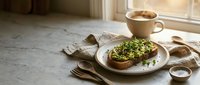 Avocado toast with microgreens on sourdough bread served on white plate with coffee cup in bright morning kitchen setting for healthy breakfast lifestyle.