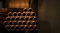 Stack of copper pipes arranged in pyramid formation with warm industrial lighting creating dramatic shadows and golden reflections on metal surfaces.