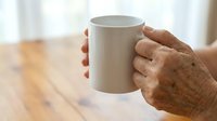 Senior adult with brown skin holding white ceramic mug on wooden table with blurred background, suitable for beverage, lifestyle, and wellness marketing materials.
