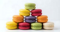 Colorful French macarons stacked in pyramid formation on white background. Assorted flavors including pistachio, lemon, raspberry, orange, chocolate cookies.