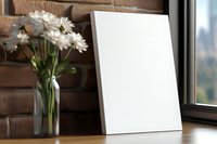 Blank white vertical frame mockup on wooden table near brick wall with daisy flowers in glass vase by window, natural lighting for artwork presentation.