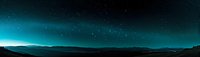 Panoramic night sky with glowing turquoise aurora borealis over dark mountain silhouettes against starry background in wilderness landscape.