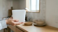 Cropped view of person holding blank white takeout cup in modern kitchen with natural light, suitable for beverage branding and food service marketing.