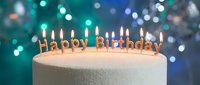 Lit golden Happy Birthday candles on white cake with bokeh teal and blue lights background, festive celebration concept.