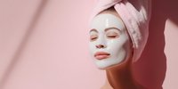 Young woman with facial clay mask and pink towel on head enjoying skincare routine against soft pink background with shadow play.