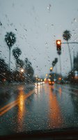 Raindrops on car windshield with blurred traffic lights and palm trees creating atmospheric bokeh effect on gloomy day, perfect for mood concepts.