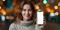 Young Caucasian woman in cozy sweater showing smartphone with blank screen against blurred bokeh lights background. Mockup for app or website design.
