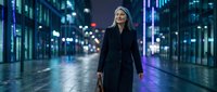Professional blonde businesswoman walking confidently through illuminated city street at night with modern buildings and neon lights creating urban atmosphere.