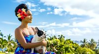 Young woman with dark hair and medium skin tone wearing colorful floral attire and red flowers in hair, holding black and white cat outdoors in tropical garden setting with palm trees and blue sky.