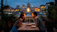 Young couple enjoying romantic dinner date on rooftop terrace with city lights background and hanging bulb lighting creating intimate evening atmosphere.